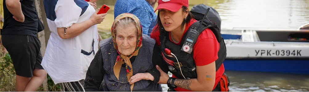 A woman, who is part of the Ukrainian Red Cross Emergency Response Team, is helping an elderly woman move away from the water. 